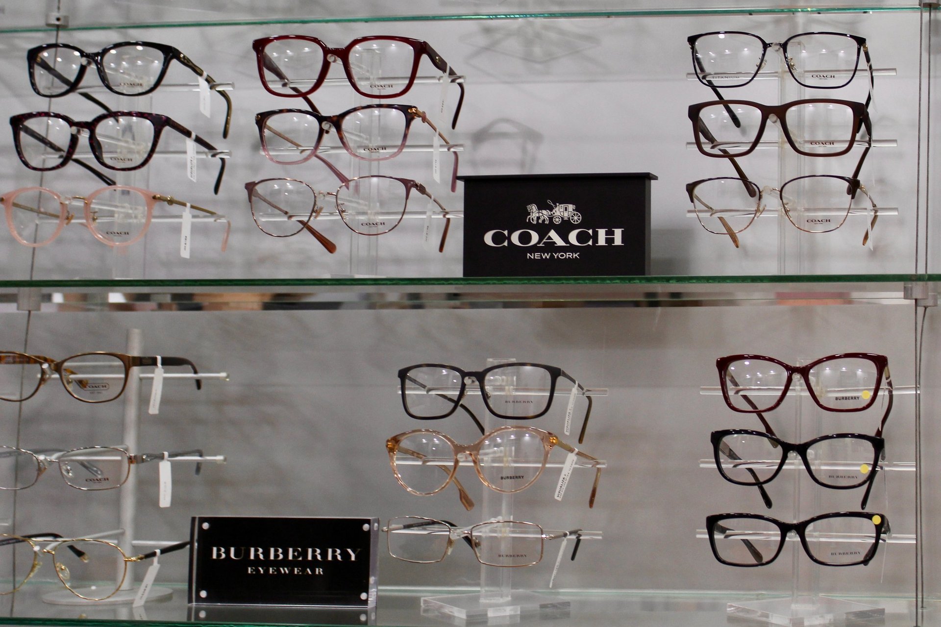 Eyeglass display case showing multiple designer frames from Coach and Burberry brands arranged on two shelves