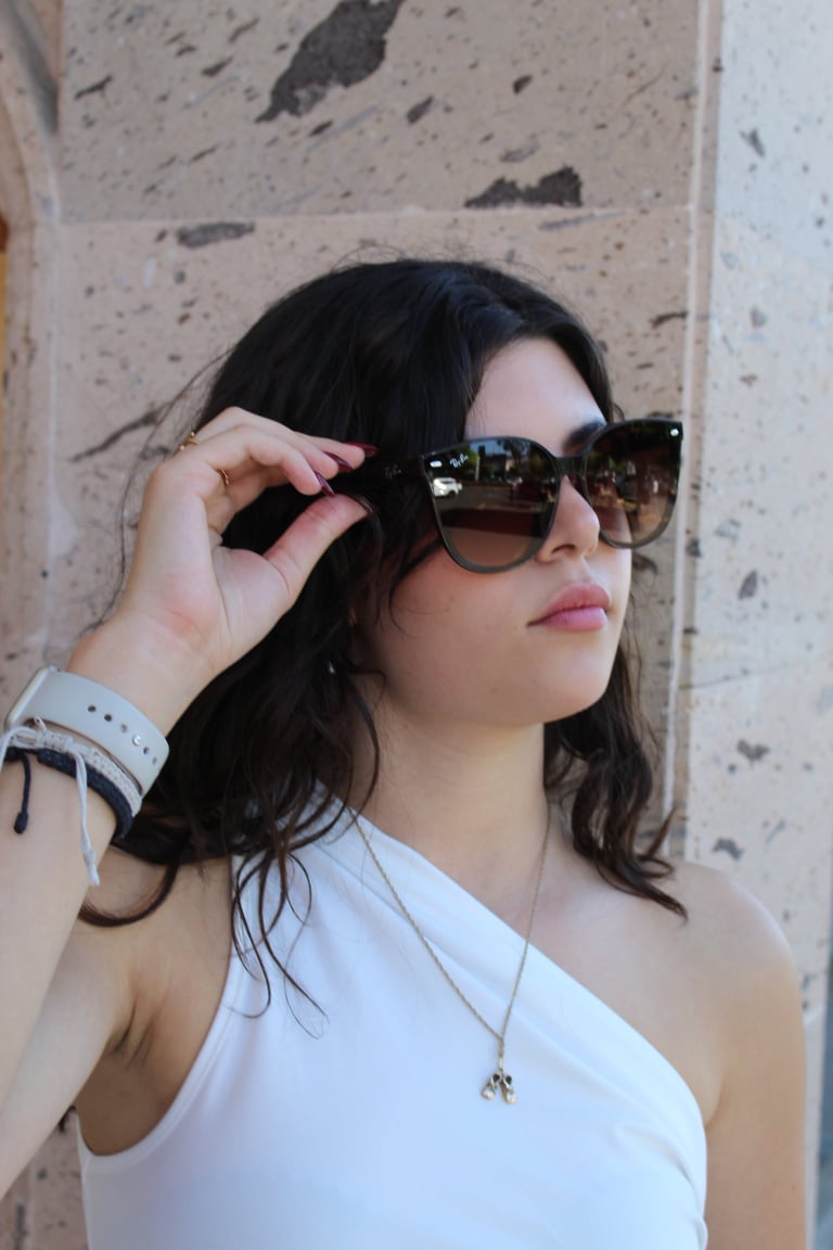 Woman with dark hair wearing white tank top and mirrored sunglasses, adjusting glasses against textured beige wall