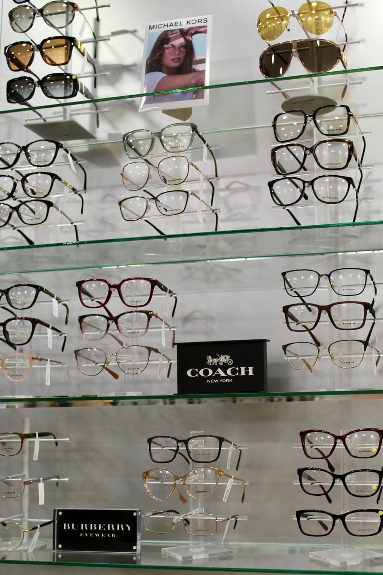 Display case with multiple eyeglasses and sunglasses arranged on glass shelves, featuring Michael Kors, Coach, and Burberry brands
