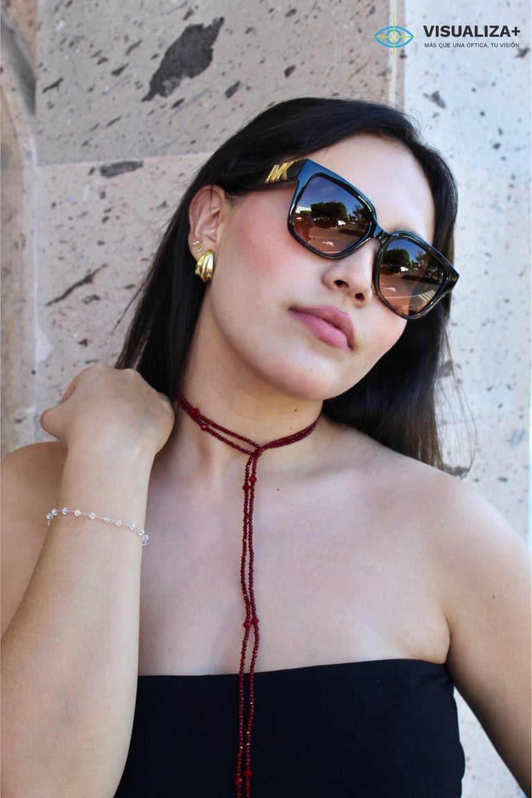 Woman wearing oversized sunglasses, gold earrings, and a beaded maroon necklace against a textured concrete wall